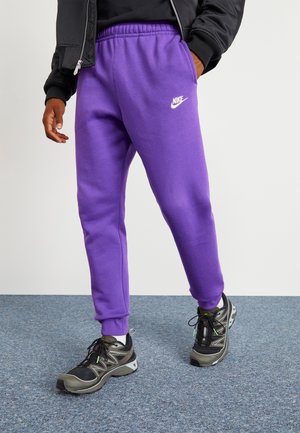 Person trägt lila Nike-Sweatpants, schwarze Jacke und schwarz-graue Sneakers und steht auf einem grauen Teppichboden.