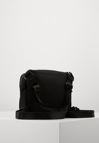 Svart axelremsväska i canvas med justerbar rem, dragkedja och enkel rektangulär design. Hållbart tyg och minimala metallbeslag.