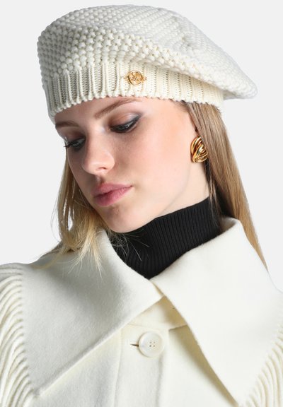 Jeune femme portant un béret en maille blanc avec détail doré, des boucles d'oreilles torsadées dorées, un col roulé noir et un manteau blanc à grand col.