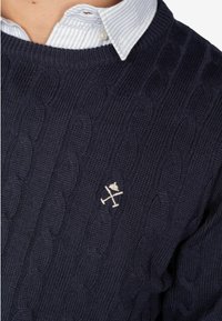 Granatowy sweter z dzianiny w sploty, z teksturowanym wzorem, z haftowanym emblematem w odcieniu kremowym po lewej stronie klatki piersiowej. Pod spodem znajduje się koszula z kołnierzykiem.