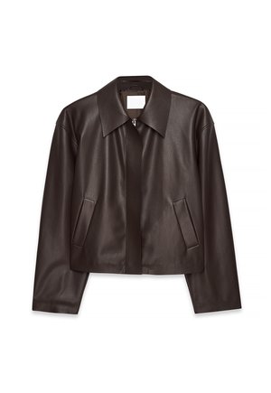 Personne marchant portant une veste en cuir marron foncé courte, une chemise rayée, un pantalon blanc ample et des sandales sur un fond clair uni.