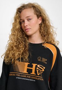 Svart sweatshirt med orange grafik, med texten "HSW" och "TJÄNAR SPELET I GENERATIONER," samt kontrasterande axeldetaljer.