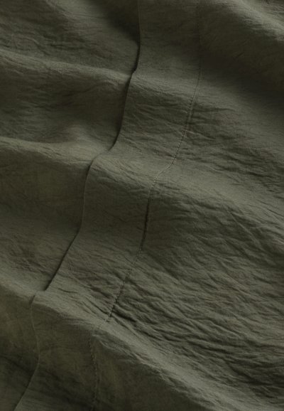 Tessuto verde oliva spiegazzato con una sottile texture, caratterizzato da linee di cucitura visibili e riflessi di luce variabili sulla superficie.