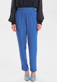 Niebieskie, prążkowane spodnie o luźnym kroju z elastycznym pasem i bocznymi rozcięciami przy kostkach, zestawione z jasnoniebieskimi butami i czarną bluzką w kropki.