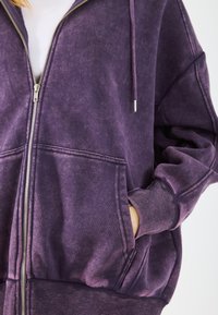 Felpa con zip viola realizzata in un tessuto morbido e testurizzato, dotata di tasche frontali, polsini a costine e cappuccio con coulisse.