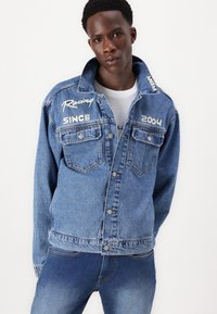 Chaqueta de mezclilla en azul medio con texto bordado "Racing" y "SINCE 2004" en los hombros y dos bolsillos en el pecho.