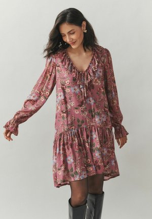 Frau in einem langen rosa Blumenkleid mit Rüschen am Ausschnitt und Saum, kombiniert mit schwarzen kniehohen Stiefeln vor einem neutralen Hintergrund.