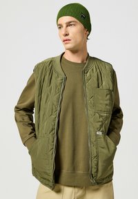 Gilet imbottito verde oliva con trapuntatura a rombi, chiusura con zip, due tasche anteriori, indossato sopra un felpa verde oliva coordinata e un berretto a coste.