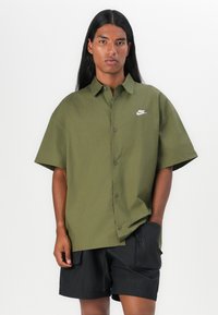 Short-sleeve button-up shirt in olive green cotton with a small white Nike logo and matching buttons, paired with black shorts.
