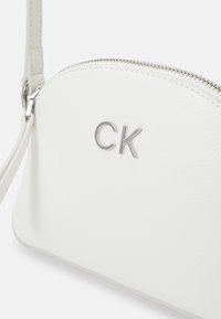 Bílá kožená crossbody taška se zaobleným tvarem, stříbrným zipem a embosovaným logem "CK" na přední straně. Hladká textura a minimalistický design.