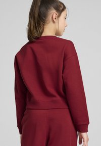 Sudadera burdeos con cuello redondo, puños y dobladillo acanalados. Tejido suave con un ajuste casual. Vista trasera con textura suave.