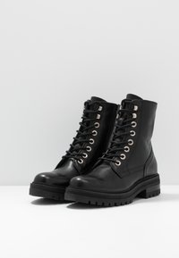 Musta keinonahkainen nilkkuri, jossa pyöreä kärki, kuusi nauhanreikää, metallikoristeita ja teksturoitu kumipohja pitävyyttä varten.