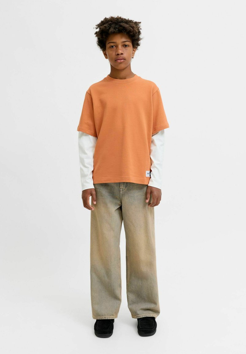 Enfant aux cheveux bouclés portant un t-shirt orange à manches courtes par-dessus un haut à manches longues blanc et un pantalon large beige avec des chaussures noires.