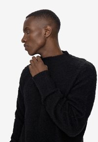 Schwarzer Wollpullover mit strukturierter, flauschiger Oberfläche. Hat einen hohen Kragen und gerippte Bündchen und wird von einer Person mit kurzen Haaren und einem Ohrring getragen.