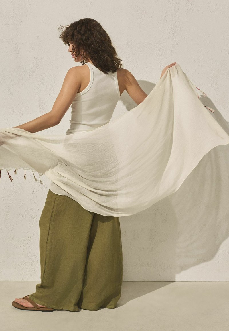Femme en débardeur blanc et pantalon large olive tenant une grande écharpe légère déployée derrière elle contre un mur texturé.