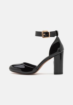 Zapato de tacón alto de charol negro con punta redondeada y correa en el tobillo con hebilla dorada.