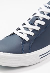 Blue leather sneaker with white laces, featuring a textured toe cap and contrasting blue stitching. White rubber sole with striped accents.