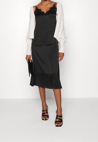 Svart satin camisole med spetsdetaljer, lager under en strukturerad vit cardigan. Svart satin kjol med utsvängd nederkant och svarta sandaletter med remmar.