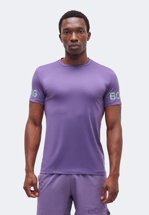 Mand med kort krøllet hår iført en tætsiddende lilla T-shirt og shortsæt med teksten "BORG" på ærmerne og shortsene.