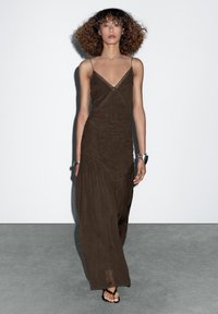 Braunes, ärmelloses Maxikleid mit V-Ausschnitt, strukturiertem Stoff und drapierter Silhouette. Getragen mit flachen schwarzen Sandalen und einem silbernen Armband.