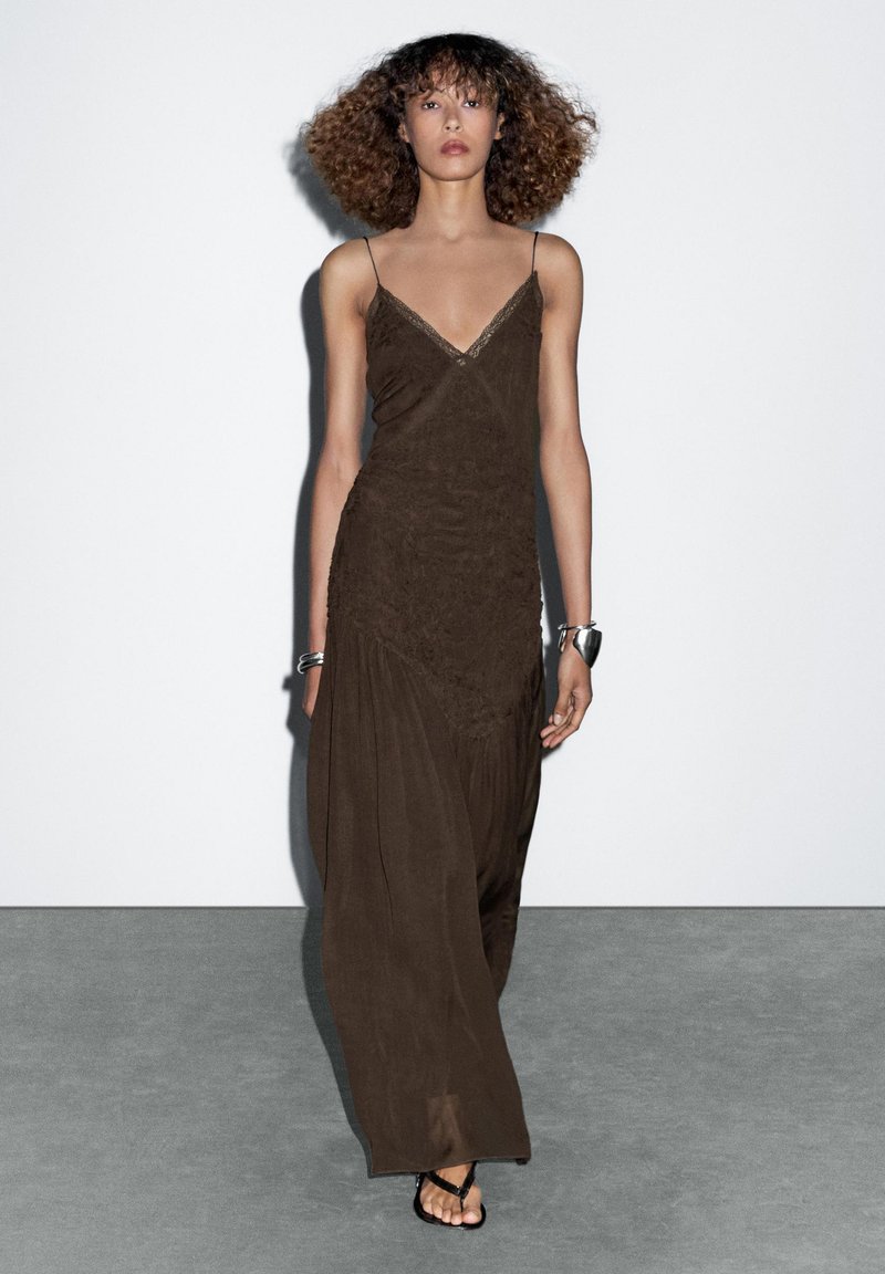 Braunes, ärmelloses Maxikleid mit V-Ausschnitt, strukturiertem Stoff und drapierter Silhouette. Getragen mit flachen schwarzen Sandalen und einem silbernen Armband.