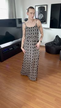 Personne avec des tatouages portant une longue robe noire et blanche à motifs, debout sur un sol en bois dans un salon avec une télévision et des fauteuils poires.