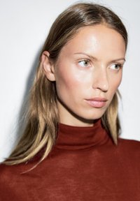 Nahaufnahme einer Person mit rotem Rollkragenpullover. Die Haut hat eine glatte Textur mit leichtem Glanz. Das Haar ist hellbraun und glatt.