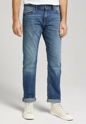 Homme portant un jean bleu retroussé et des baskets blanches montantes, debout devant un fond uni.