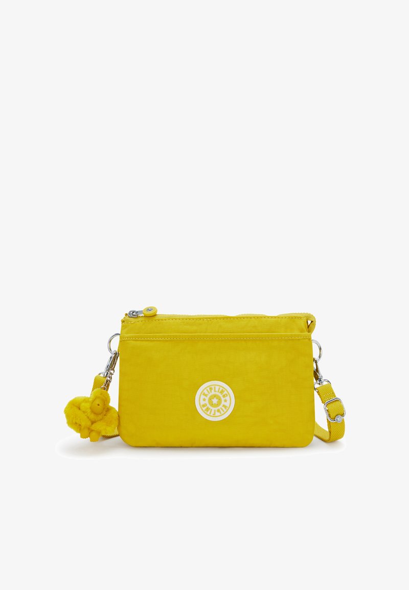 Sac bandoulière en nylon jaune avec fermeture éclair, orné d'un logo rond et d'un porte-clés duveteux. Texture lisse, forme rectangulaire compacte.