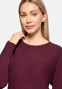 Bordeauxrode gebreide trui met een ronde hals, geribbelde manchetten en een gladde textuur, gedragen door een persoon met steil blond haar, hand op de nek.