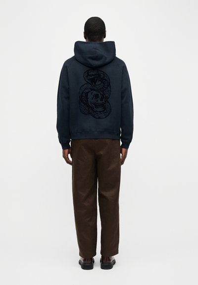 Άνδρας με μπλε-σκούρο φούτερ με κουκούλα που έχει μαύρο γραφικό φίδι και κρανίο στην πλάτη, καφέ παντελόνι και σκούρα παπούτσια, στέκεται γυρισμένος με την πλάτη σε λευκό φόντο.