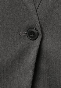 Grijze stof met een fijne weving, voorzien van een ronde zwarte knoop en subtiele stikdetails. Een klassiek, gestructureerd jasje-element.