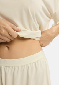 Jasno beżowy zestaw odzieży domowej o miękkiej teksturze. Bluzka ma luźny krój; spodnie z elastyczną talią z logo "ESOTIQ" na pasku.