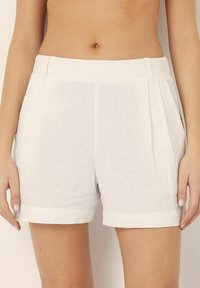 Pantalones cortos de lino blanco con un ajuste relajado, que cuentan con pliegues en la parte delantera, dobladillos enrollados y una textura suave.