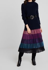 Maglione lavorato a maglia blu navy abbinato a una gonna plissettata multicolore con righe orizzontali in viola, turchese e nero. Dettaglio di cintura nera.