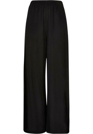 Femme portant un pantalon noir ample avec taille élastique et des sandales à plateforme noires, debout de profil sur un fond uni.