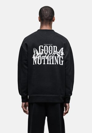 Schwarzer Pullover mit grafischem Text auf dem Rücken, der die Worte "PRÄSENTIERT VON GOOD FOR NOTHING" und Schmetterlingsillustrationen zeigt.