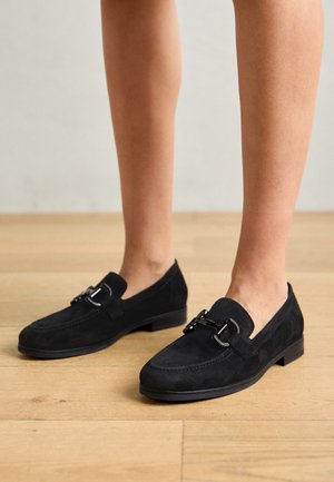Sorte loafers i ruskind med flad sål, der har karakteristisk sølvkæde over vristen og rundet tådesign.