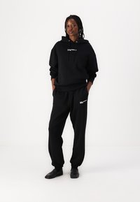 Black hoodie with front pocket and drawstring, paired with matching black sweatpants featuring logo accents. Material appears soft and relaxed fit.