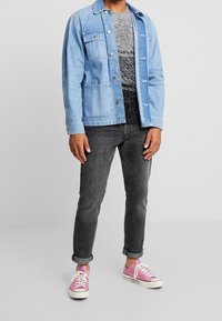 Veste en denim bleu clair avec deux poches poitrine, portée sur une chemise noire et grise à motifs. Jean gris foncé retroussé et baskets basses roses.