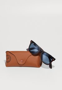 Sluneční brýle Ray-Ban s tmavými skly, obdélníkovým tvarem a silnými černými obrubami; v kombinaci s texturovaným hnědým koženým pouzdrem, které má uzávěr na knoflík.