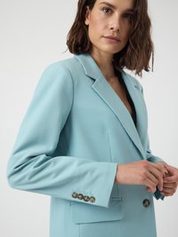 Woman adjusting light blue blazer with dark buttons, wearing black top underneath, against a plain light background.