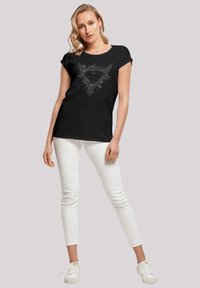 Black t-shirt featuring a graphic design with a central logo and wing accents. Short sleeves, relaxed fit, paired with white skinny jeans.