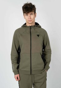 Felpa con zip di colore verde oliva, realizzata in materiale morbido, con pannelli laterali neri, cappuccio e un piccolo logo sul petto sinistro.