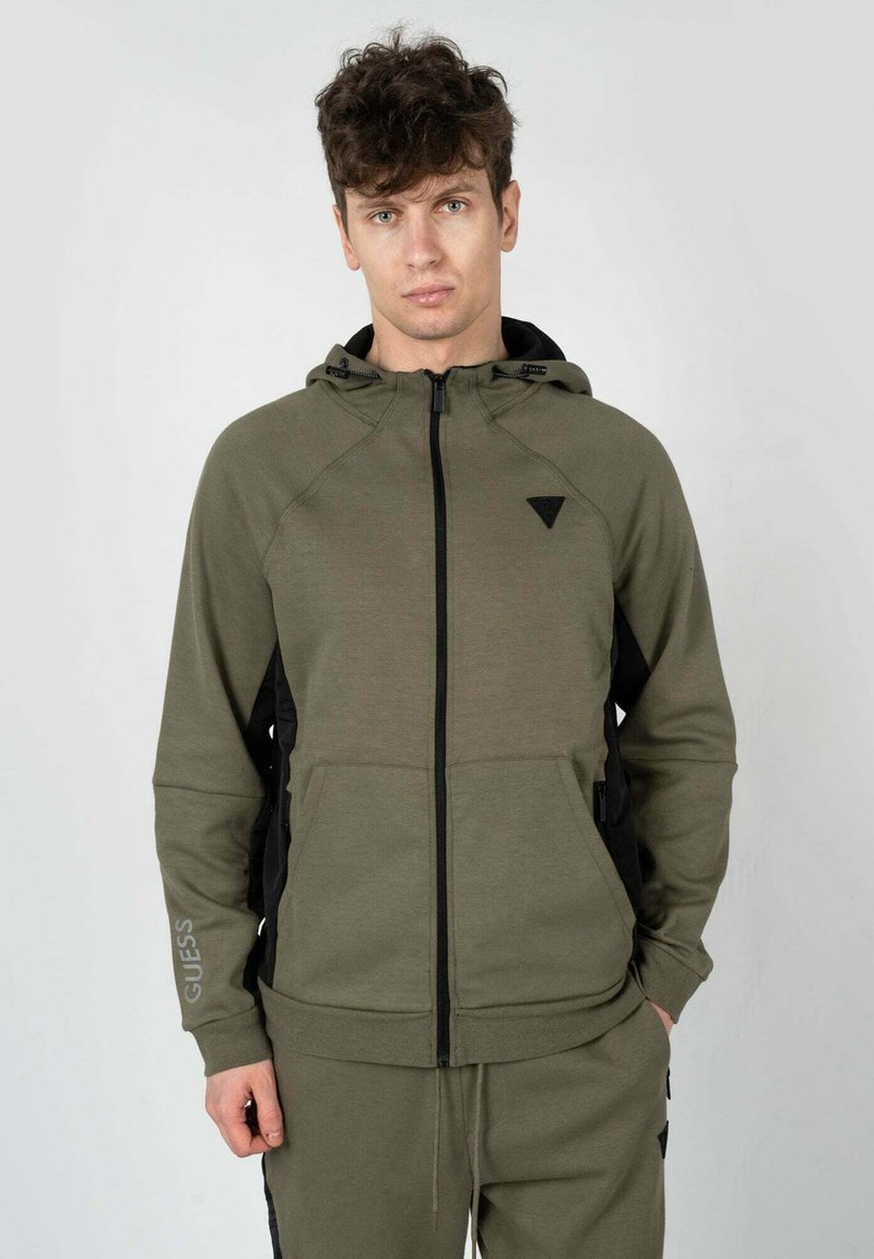Felpa con zip di colore verde oliva, realizzata in materiale morbido, con pannelli laterali neri, cappuccio e un piccolo logo sul petto sinistro.
