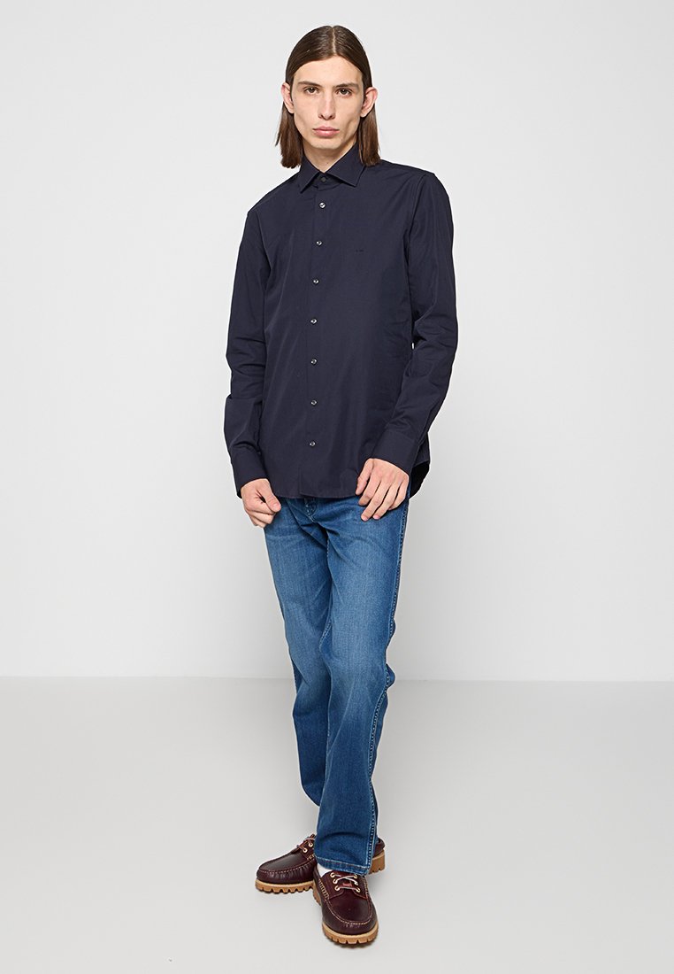 Giovane uomo in piedi che indossa una camicia blu scura con bottoni, jeans blu e scarpe di pelle marroni su uno sfondo grigio chiaro.