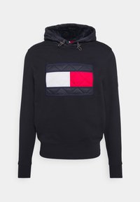 Granatowy hoodie z pikowanym kapturem na sznurku oraz dużą prostokątną łatą z białymi i czerwonymi blokami kolorów z przodu.