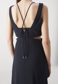 Dos d'une robe noire sans manches avec décolleté plongeant, découpes sur les côtés et fines bretelles croisées nouées en un nœud au centre du dos.