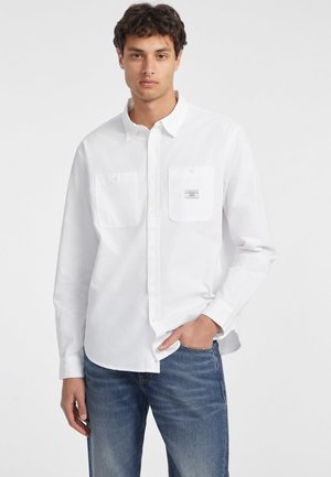 Valge pika varrukaga nööbiga särk, millel on kaks eestpoolse taskut, krae ja sildiga vasakul taskul, koos helesiniste teksadega.