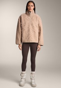 Džemper iz flisa v bež barvi z teksturiranim cik-cak vzorcem, kombiniran s rjavimi legicami in belimi škornji z vezalkami ter mehkim ovratnikom.